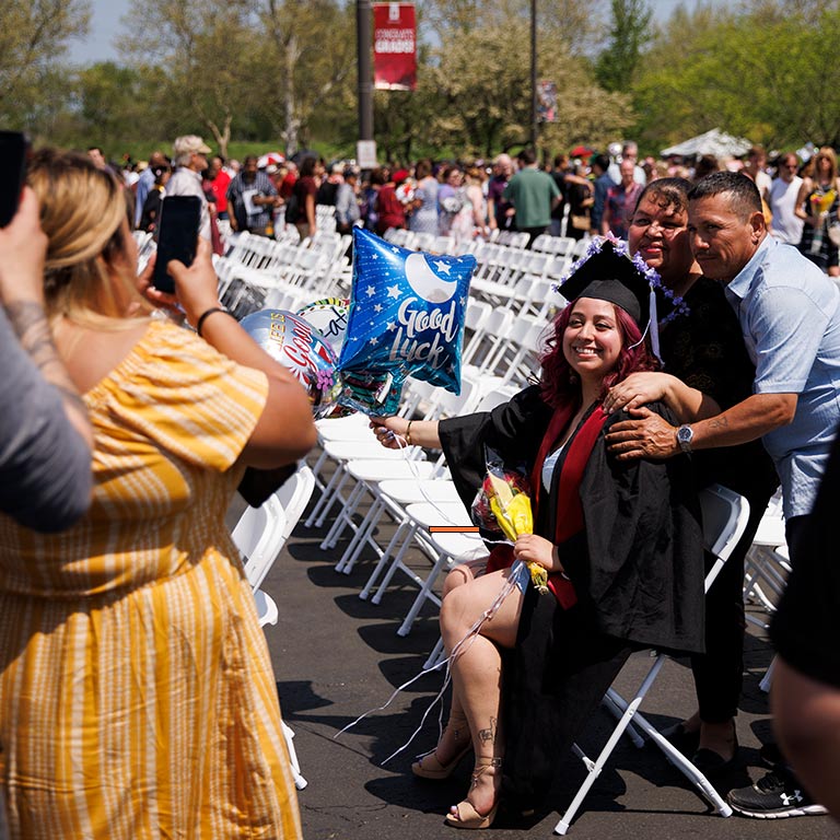 A graduate sits at an outdoor chair with friends and family nearby, all posing for a photo