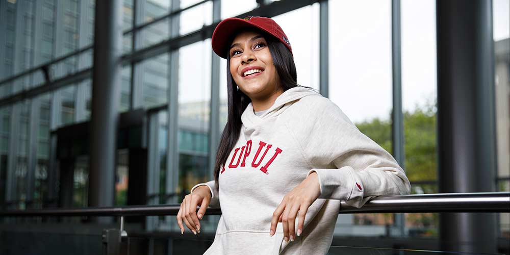 Female student leaning against a railing with an IUPUI sweatshirt and red hat.