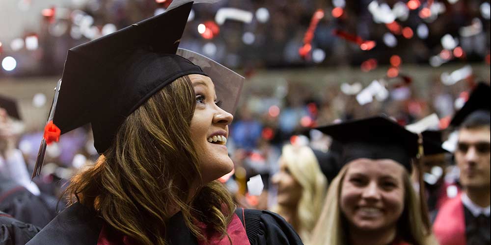 A female graduate in cap in robe smiling with other graduates in the background.